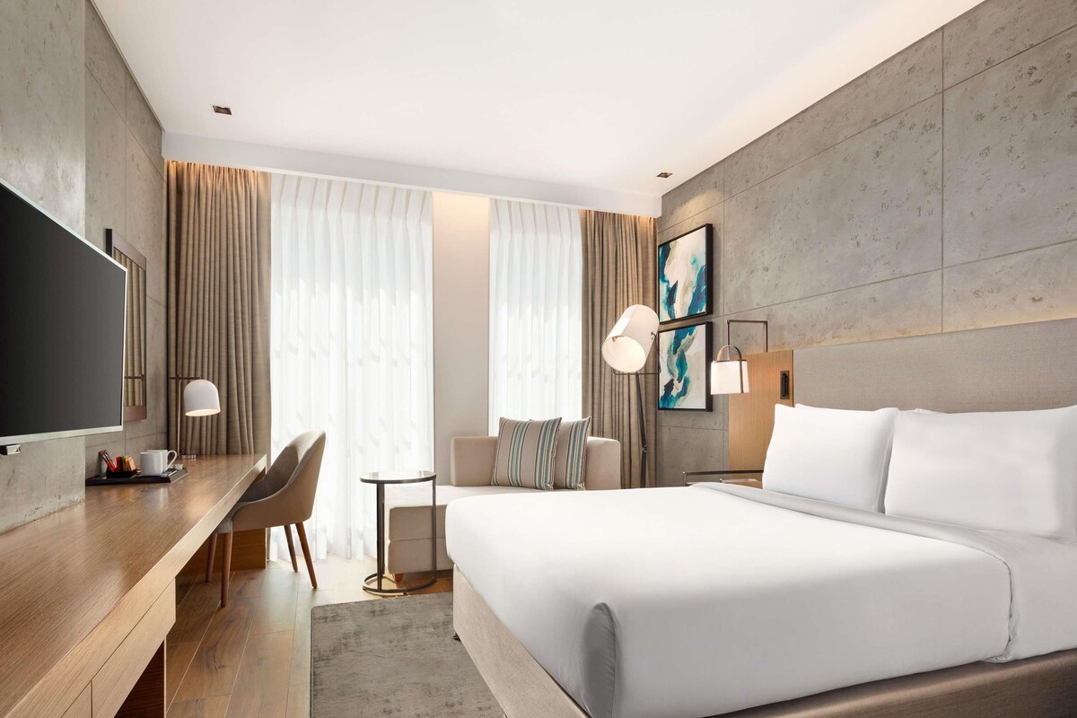 A modern hotel room with a large bed, a desk with a chair, a TV, and a small table.