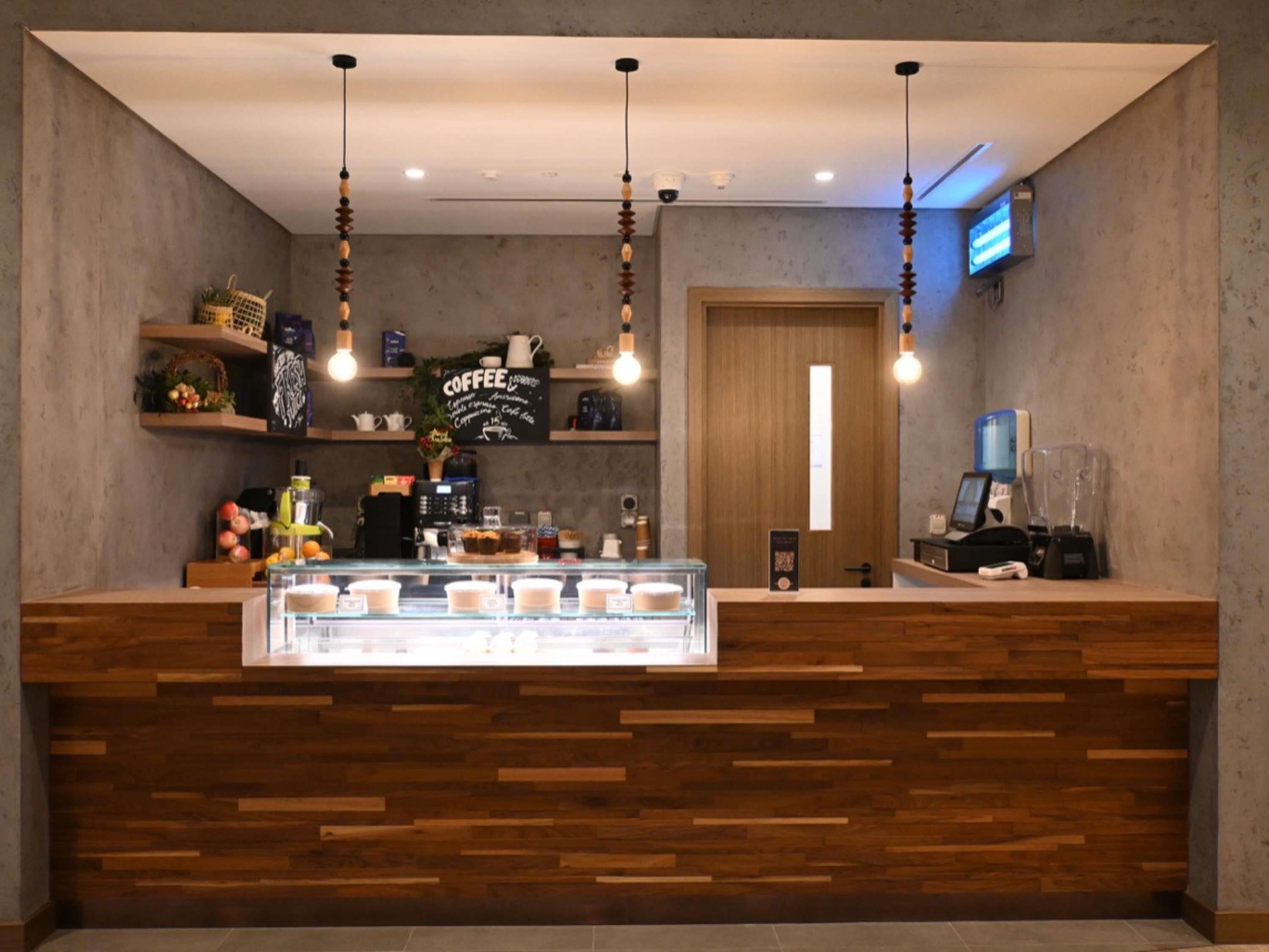Interior de una cafetería con mostrador de madera, luces colgantes y un letrero de café en la pared.