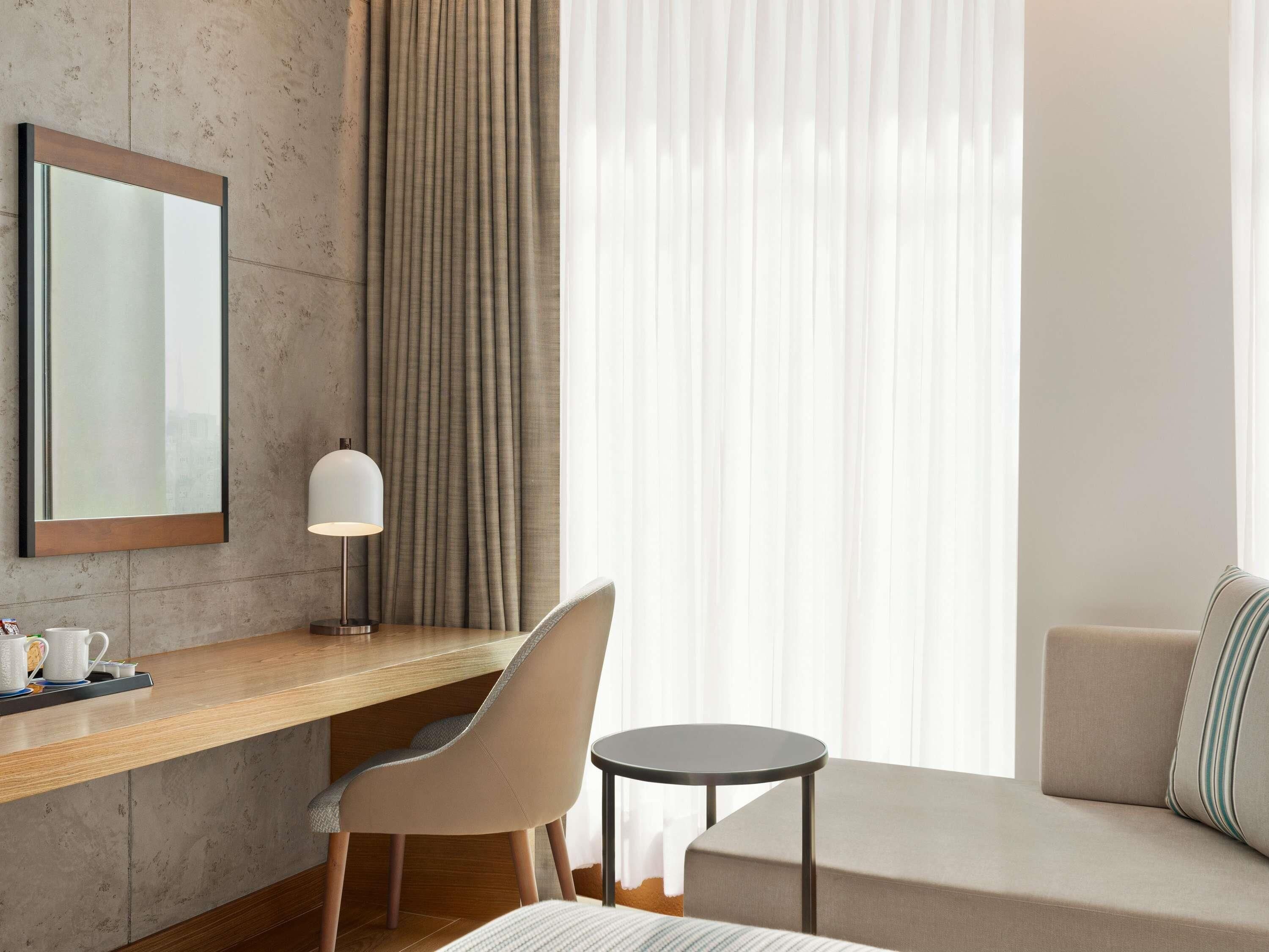 Ein Hotelzimmer mit einem Holztisch, einem Stuhl, einem runden Tisch, einer Couch und einem großen Fenster mit Vorhängen.