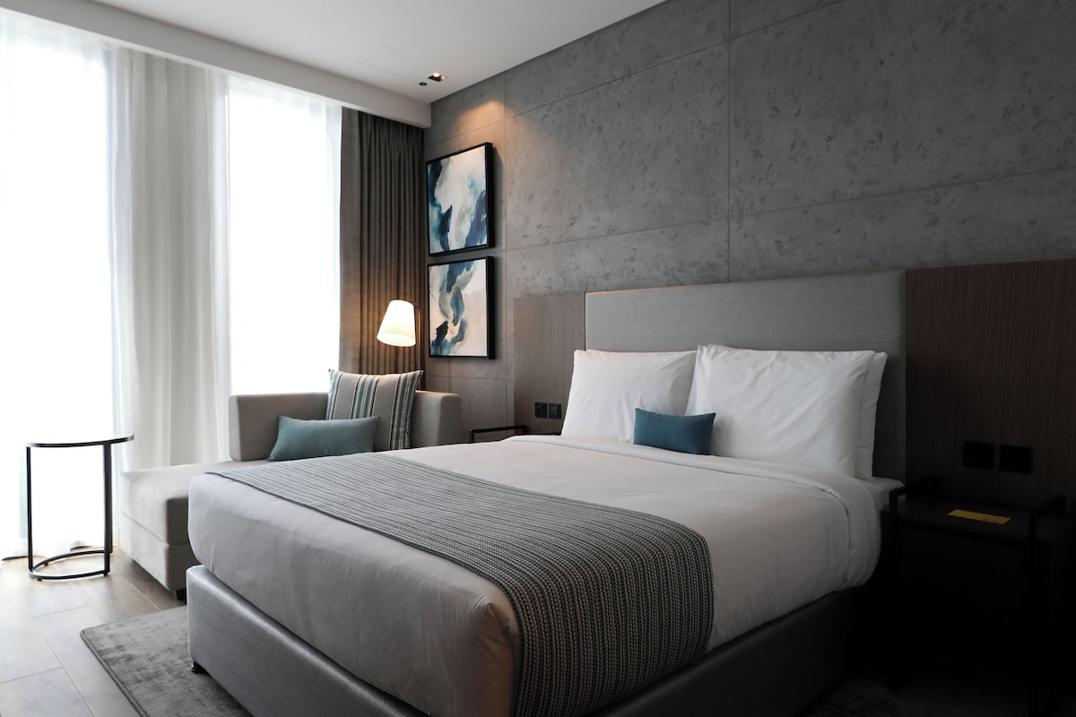 A modern hotel room with a large bed, a nightstand, a small table, and a view of the outdoors.