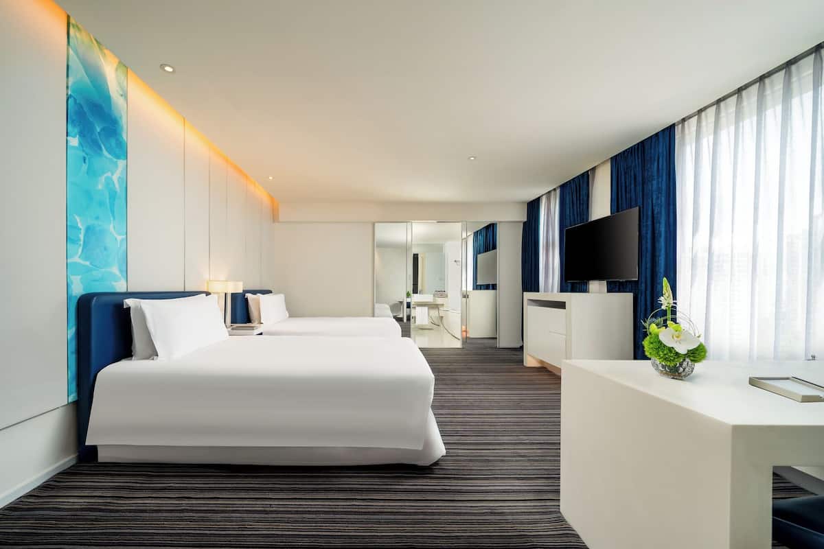 Ein modernes Hotelzimmer mit einem großen Bett, einem Schreibtisch, einem Flachbildfernseher und einem durch eine Glastrennwand einsehbarer Badezimmer.