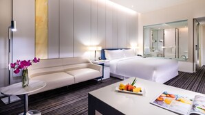 Een moderne hotelkamer met een bed, een bank, een klein tafeltje met fruit en een tijdschrift, een nachtkastje met een lamp en een grote spiegel.