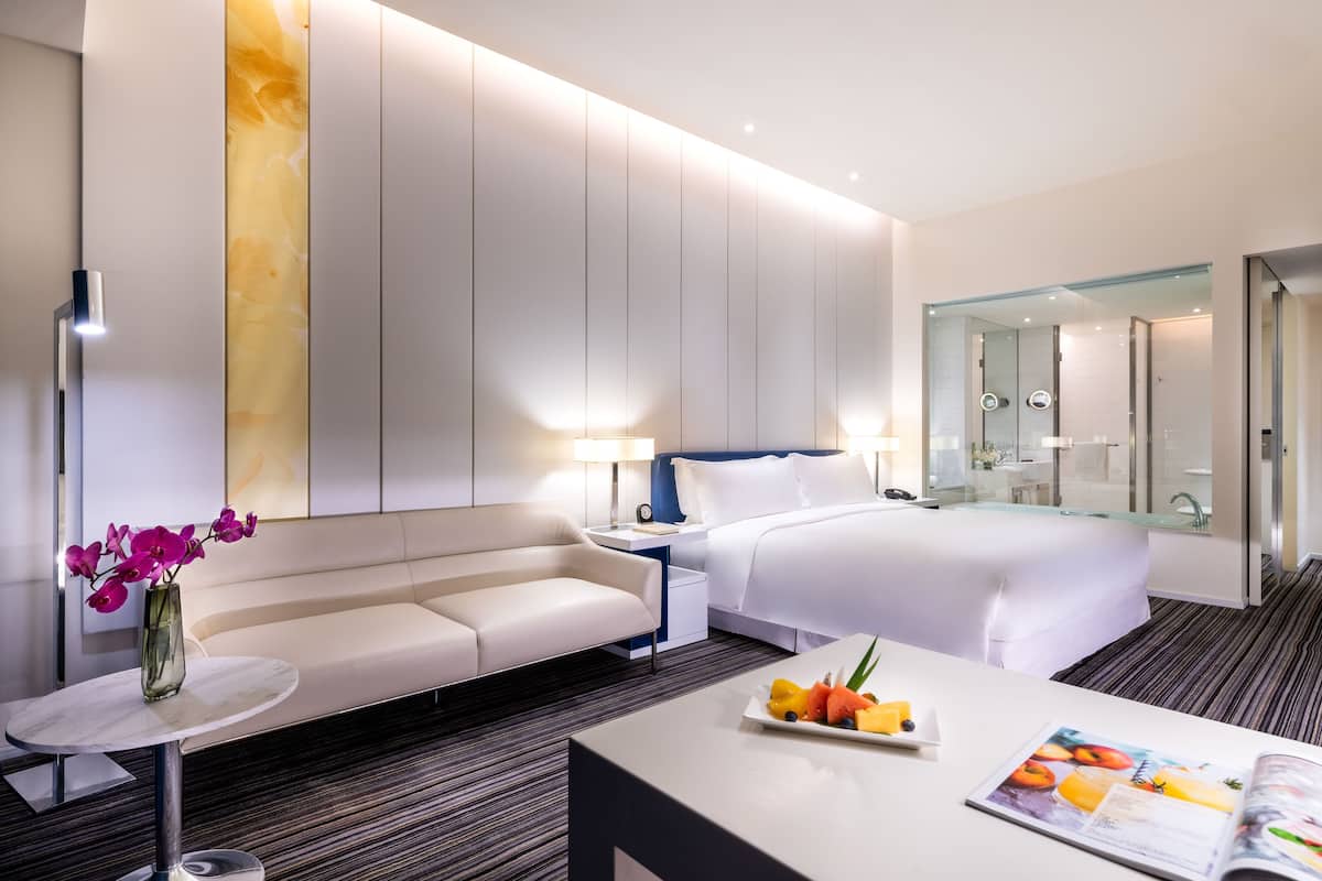 Ein modernes Hotelzimmer mit Bett, Sofa, kleinem Tisch mit Früchten und Zeitschrift, Nachttisch mit Lampe und großem Spiegel.