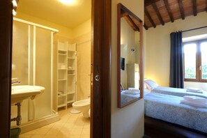Ein Badezimmer mit Toilette, Waschbecken und Dusche. Zwei Betten mit Handtüchern sind vorhanden.