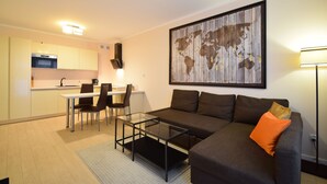 A modern living area with a glass coffee table, a sofa with an orange pillow, a dining table with chairs, and a kitchen in the background. - RentPlanet - Bliżej Morza (Kolobrzeg)