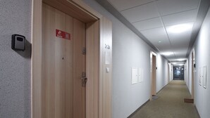 A long, narrow hallway with wooden doors on one side and white walls on the other. There are signs on the doors, a fire alarm on the wall, and a carpeted floor. - RentPlanet - Bliżej Morza (Kolobrzeg)