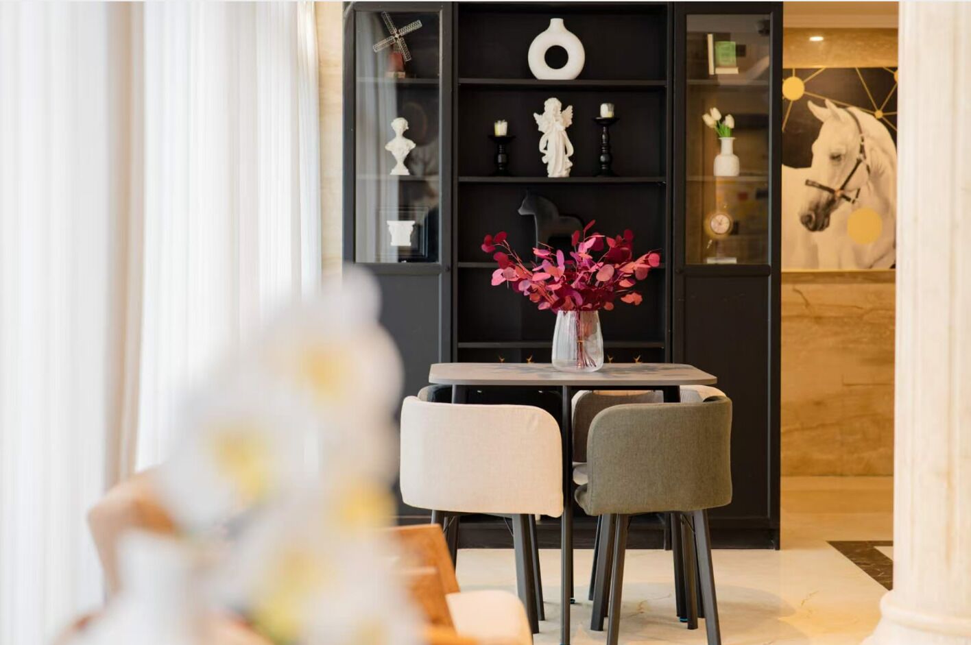 A dining area with a table and chairs, a vase with flowers, and a black shelving unit with decorative items.