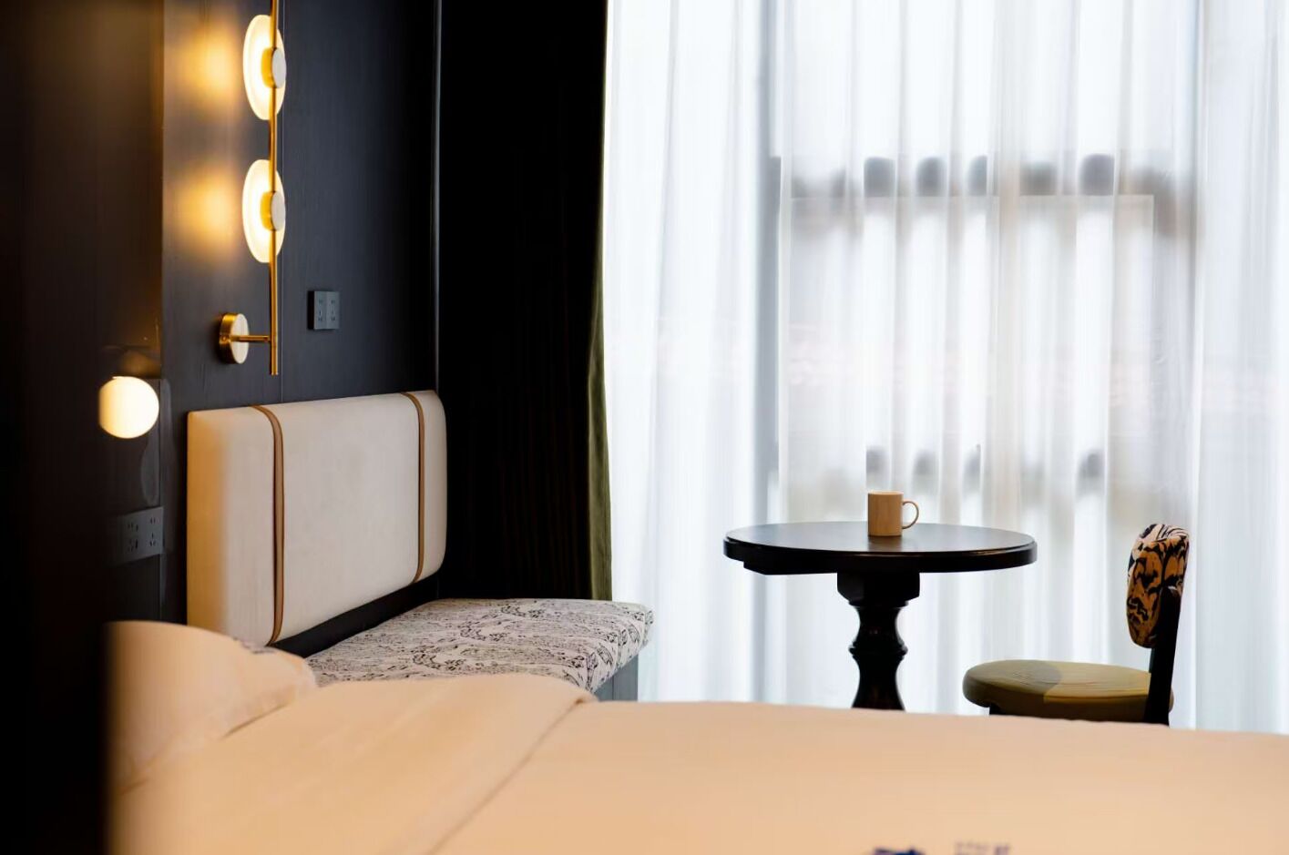 Een hotelkamer met een bed, nachtkastje met een kopje, een stoel en een wandlamp.