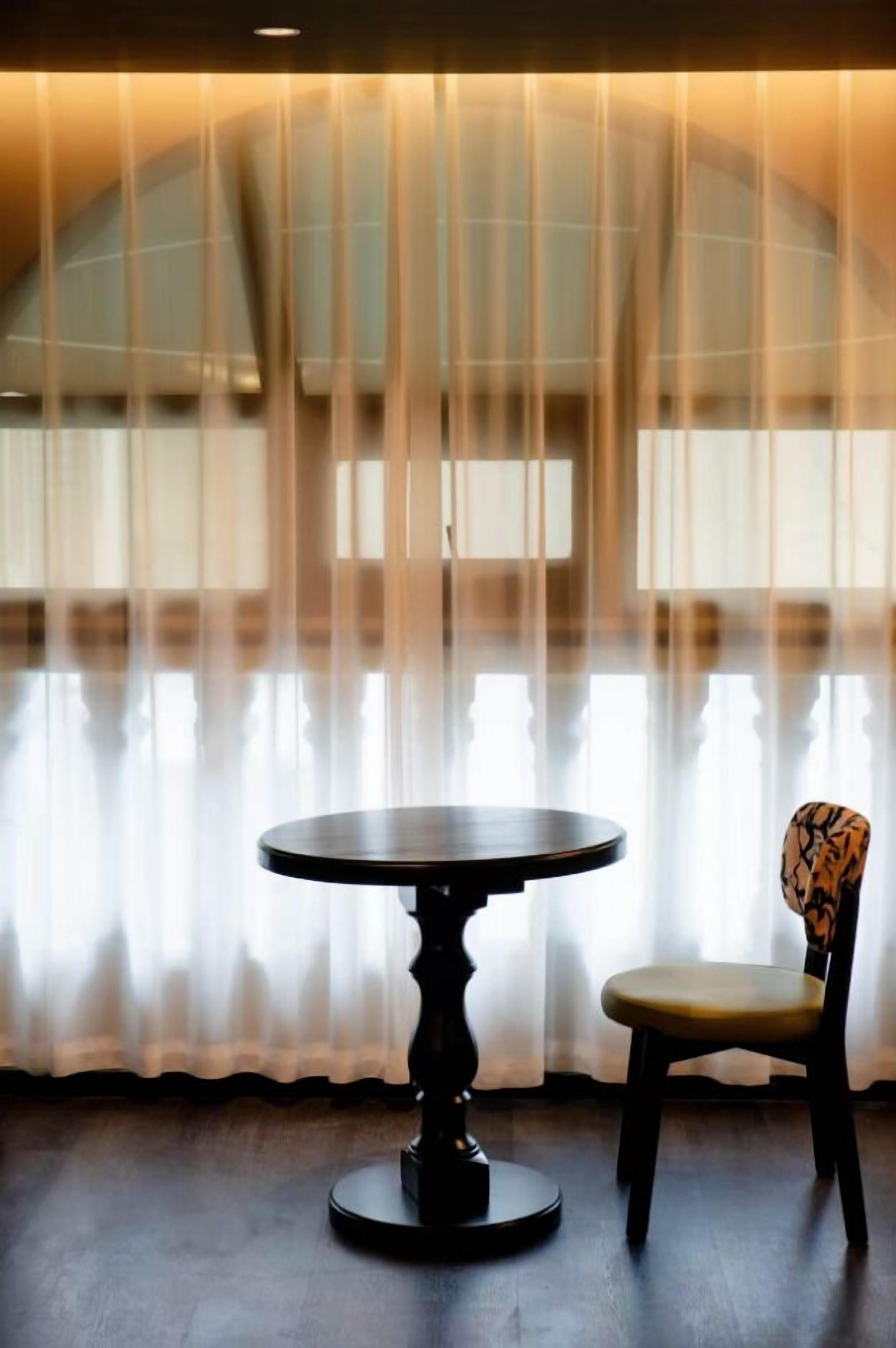 Ein Zimmer mit einem runden Tisch und einem Stuhl, einem großen Fenster mit durchscheinenden Vorhängen und einer gewölbten Decke.