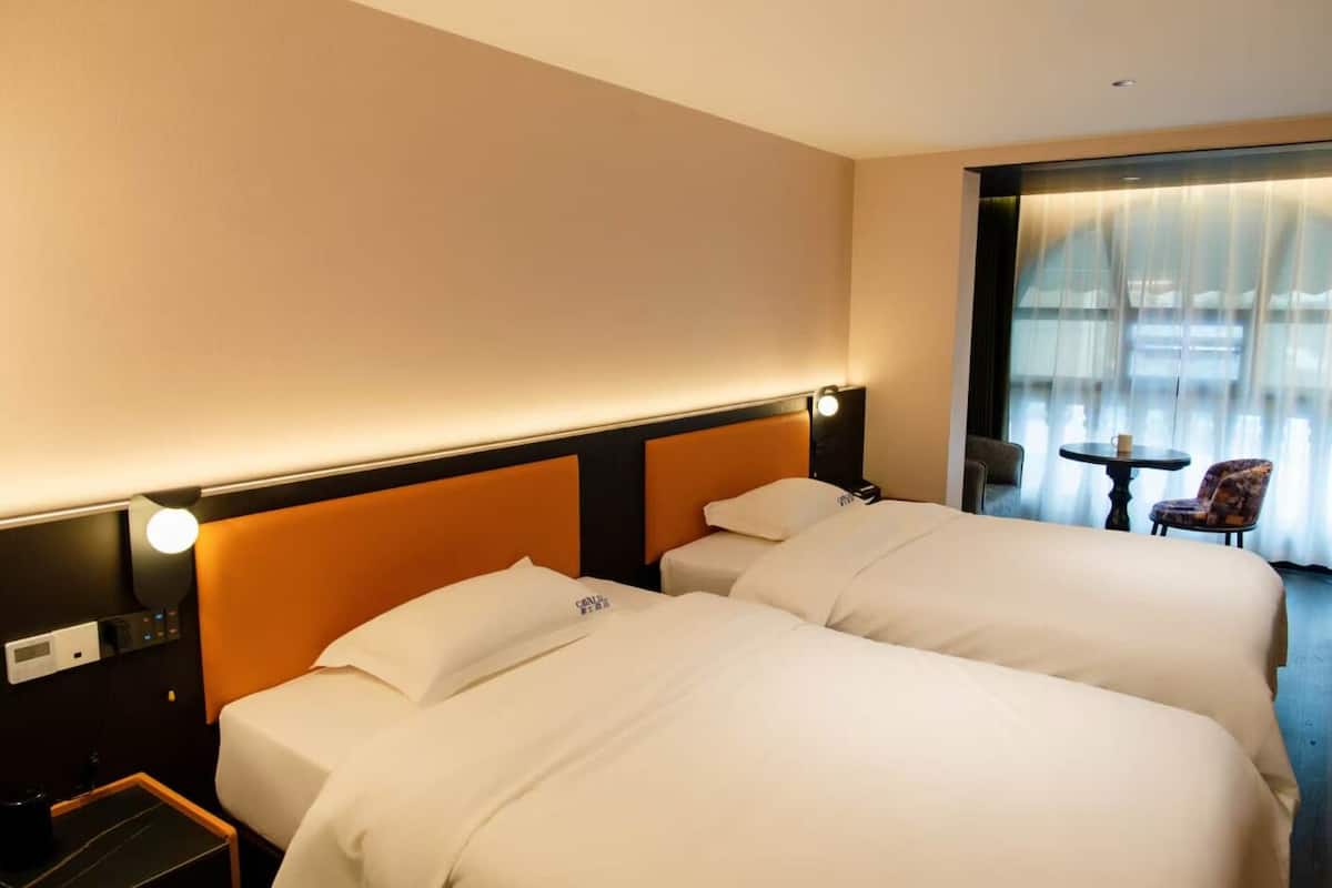 A hotel room with two beds, a nightstand, a lamp, a chair, and a table with a cup.
