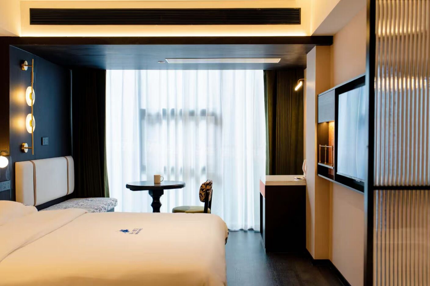 Ett hotellrum med en stor säng, ett litet bord, en stol, en tv och ett fönster med gardiner.