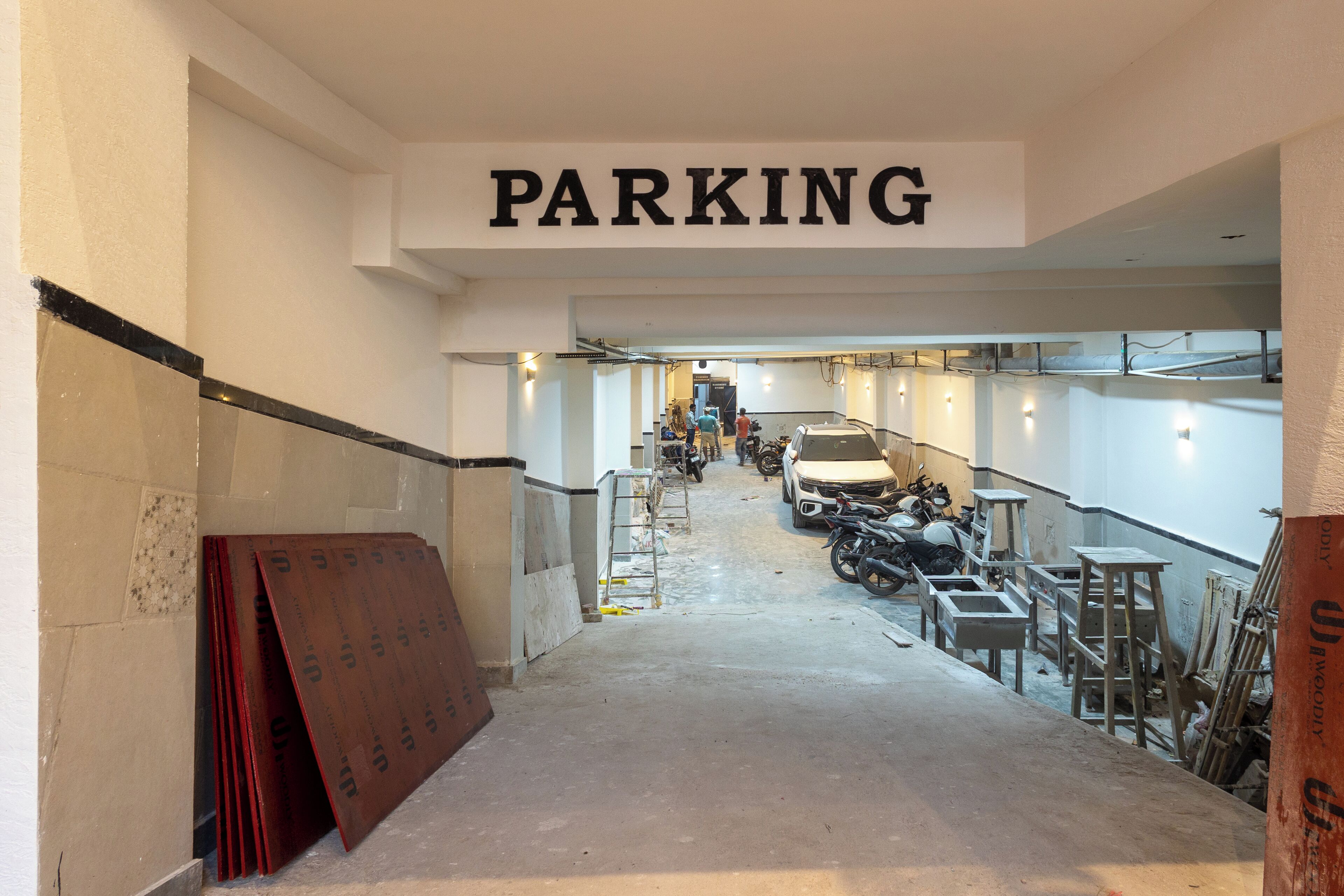 Un pasillo estrecho con un letrero de 'PARKING' encima, que conduce a una zona de estacionamiento con varias motocicletas y un coche blanco.