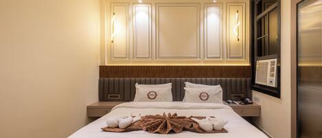 A hotel room with a large bed, a nightstand, a wall-mounted TV, and a window with blinds.