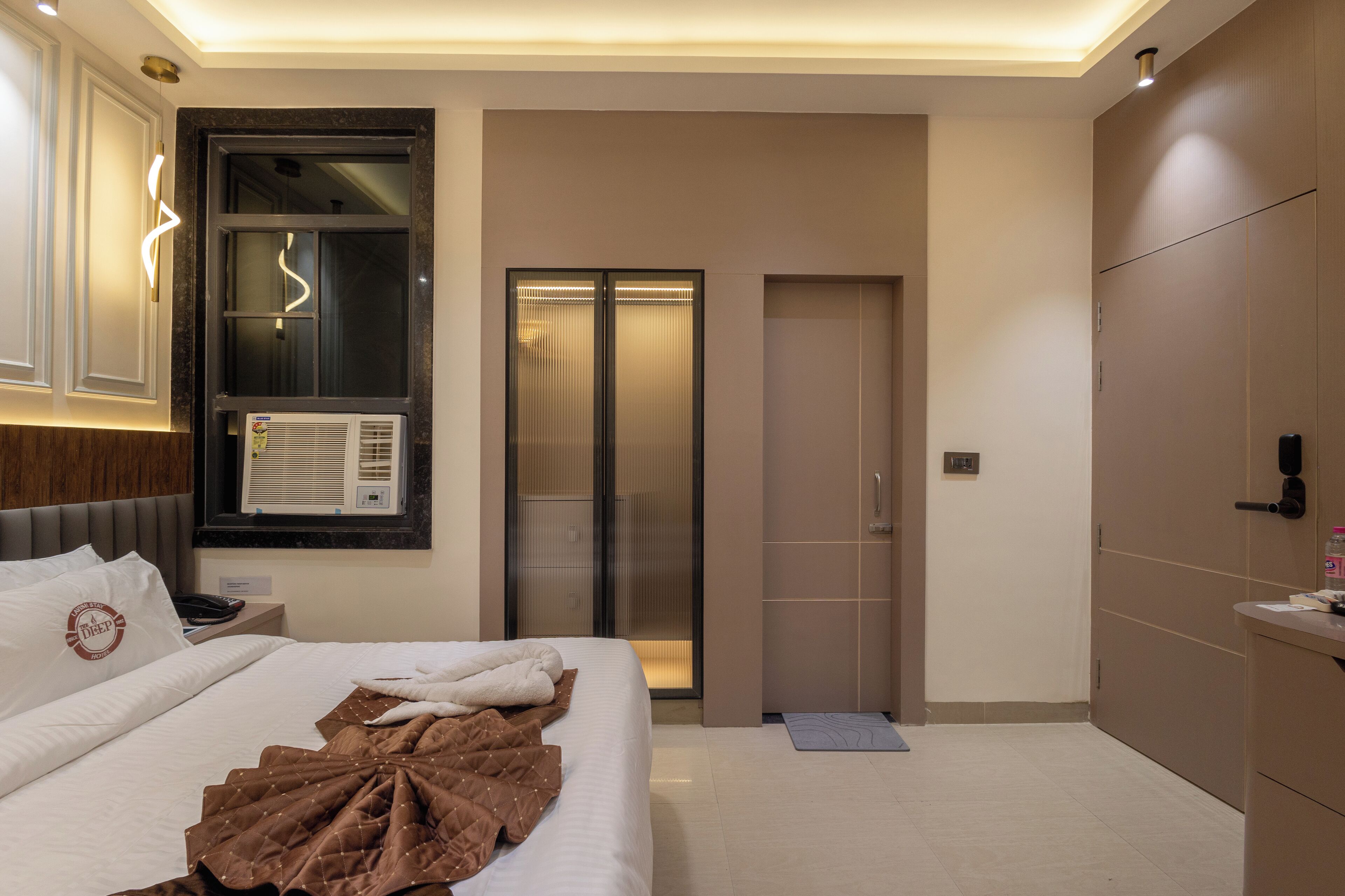 A modern hotel room with a neatly made bed, a large window with blinds, a built-in air conditioning unit, and a sliding door leading to a bathroom.