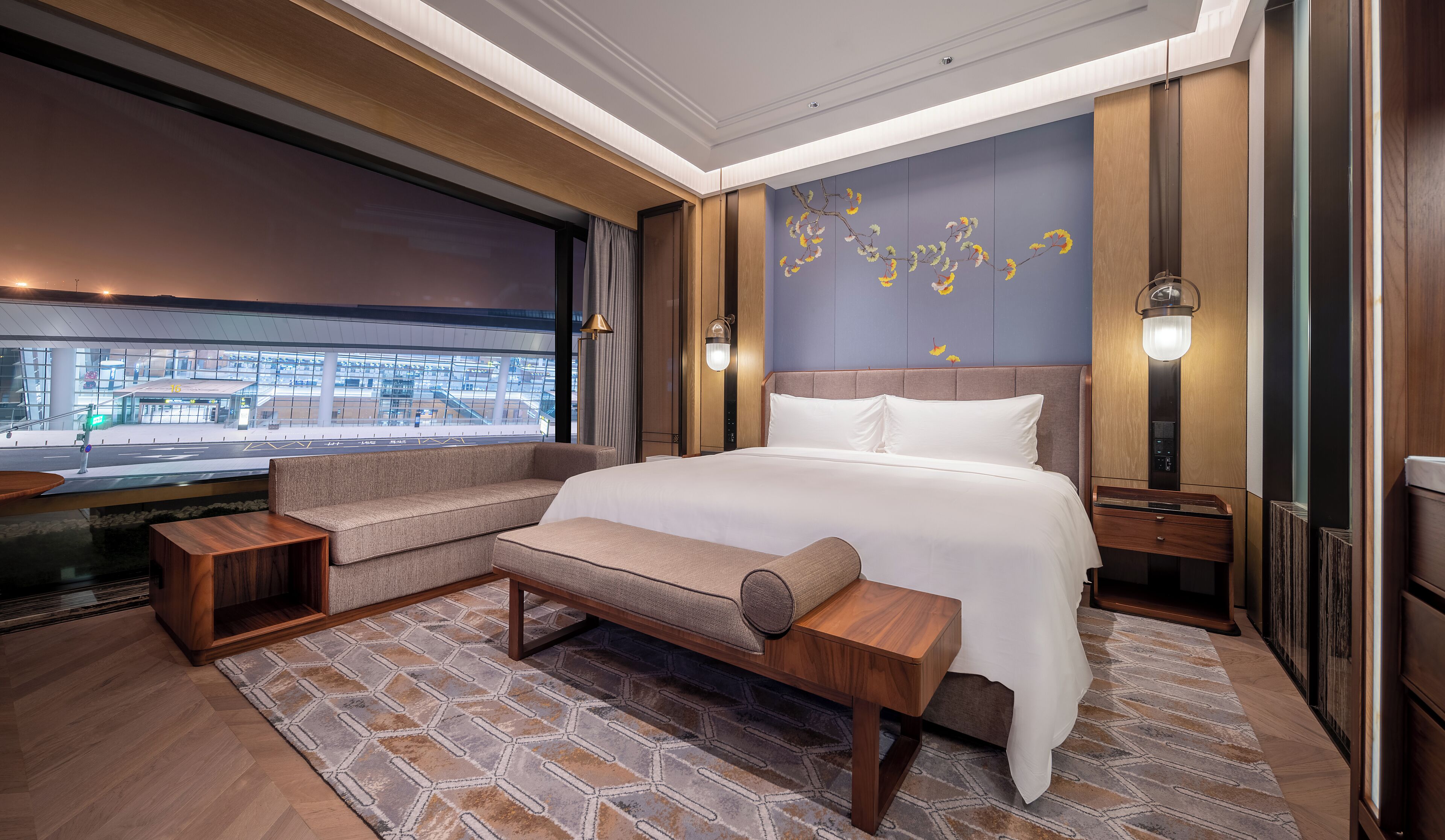 A modern hotel room with a large bed, a sofa, a bench, and a view of the cityscape through large windows.