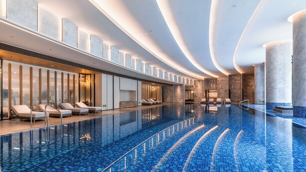 Piscina interior moderna com chão de azulejos azuis e brilhantes, espreguiçadeiras ao longo da borda e um design de teto curvo e elegante.