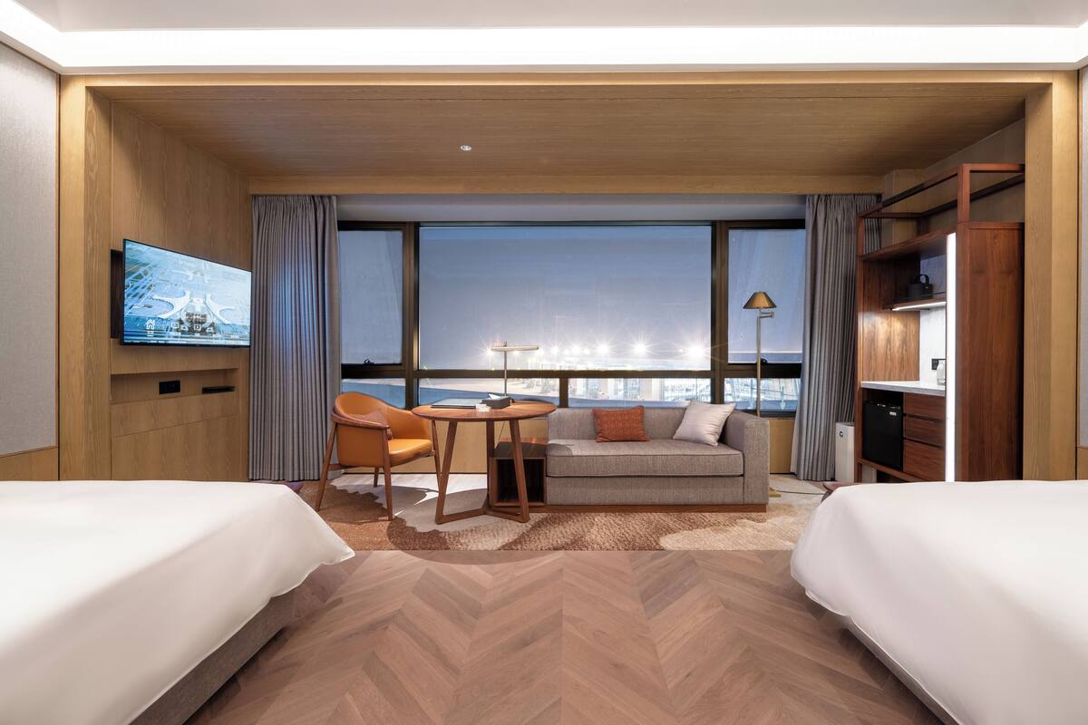 Ett hotellrum med två sängar, en soffa, ett litet bord, en TV och utsikt över stadsbilden.