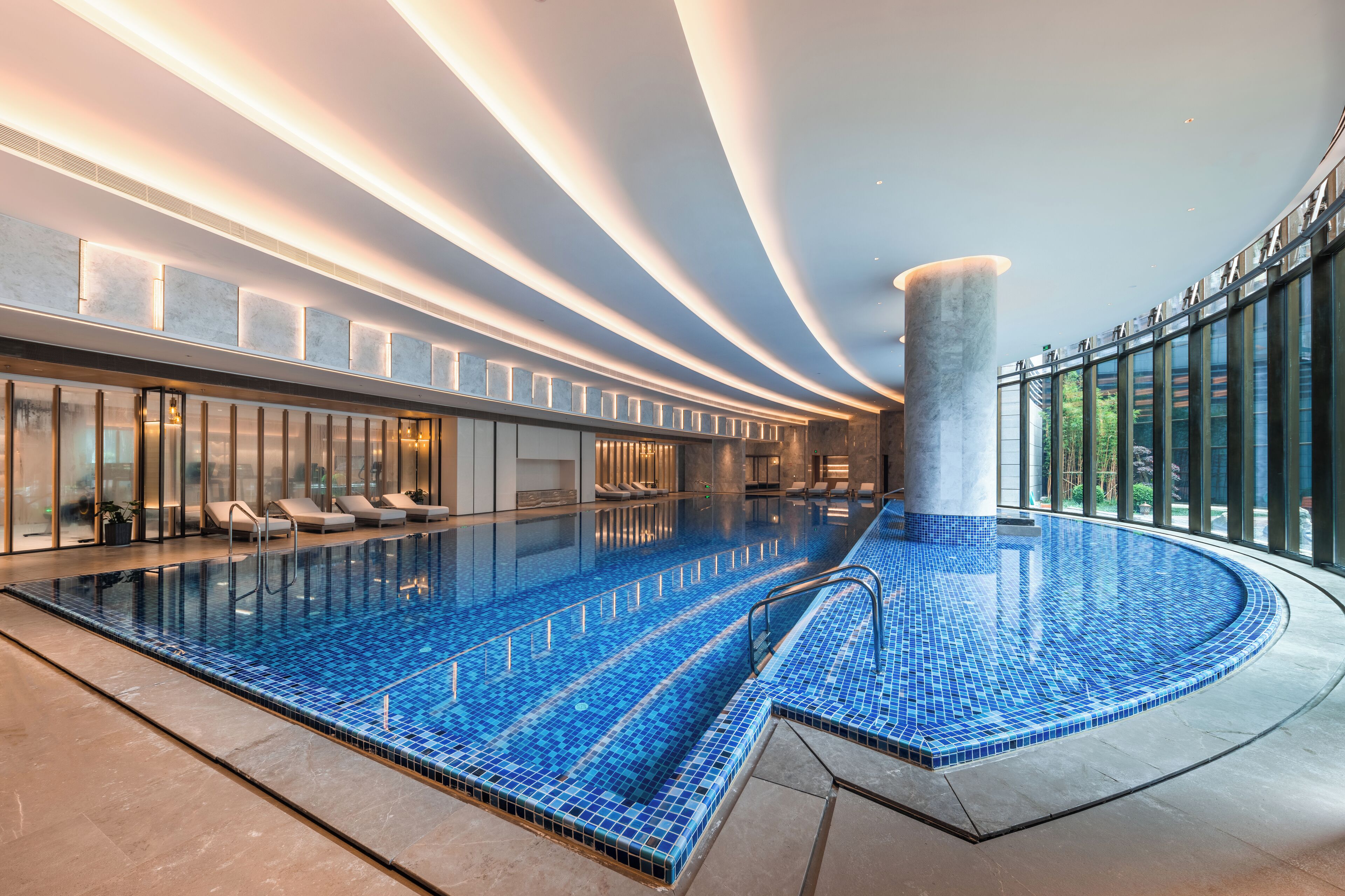 A modern indoor swimming pool with a curved design, surrounded by lounge areas and large windows.