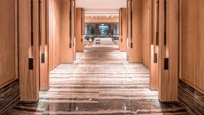 Corredor com painéis de madeira e chão de mármore, que leva a uma área de recepção com portas de vidro.