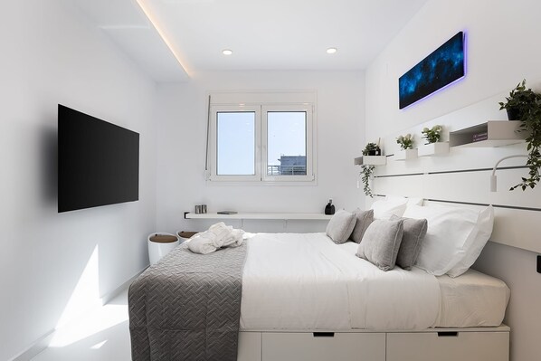 A modern bedroom with a bed, a flat-screen TV, a window, and a wall-mounted shelf with plants.