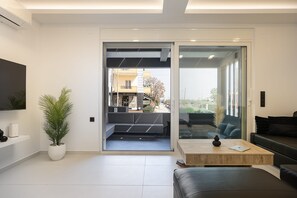 A modern living room with a sliding glass door leading to an outdoor area, a flat-screen TV mounted on the wall, a potted plant, and a leather sofa.