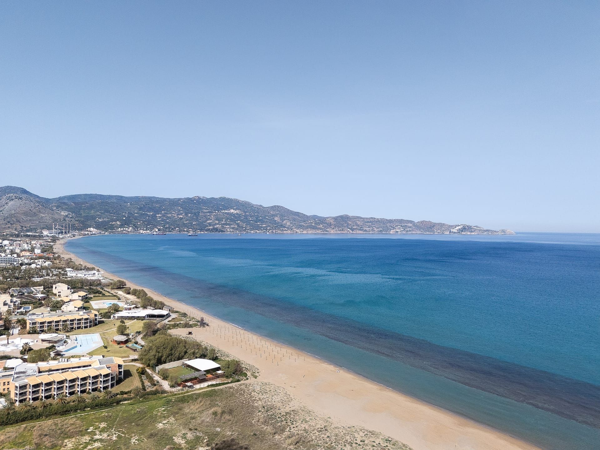 A coastal view with a beach, buildings, and a hilly landscape in the background.
