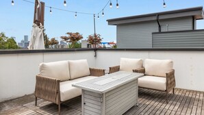 An outdoor seating area with a table and two sofas, string lights overhead, and a cityscape in the background.