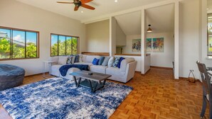 Un salon spacieux avec un grand canapé, une table basse et un ventilateur de plafond. De grandes fenêtres offrent une vue sur la verdure environnante.