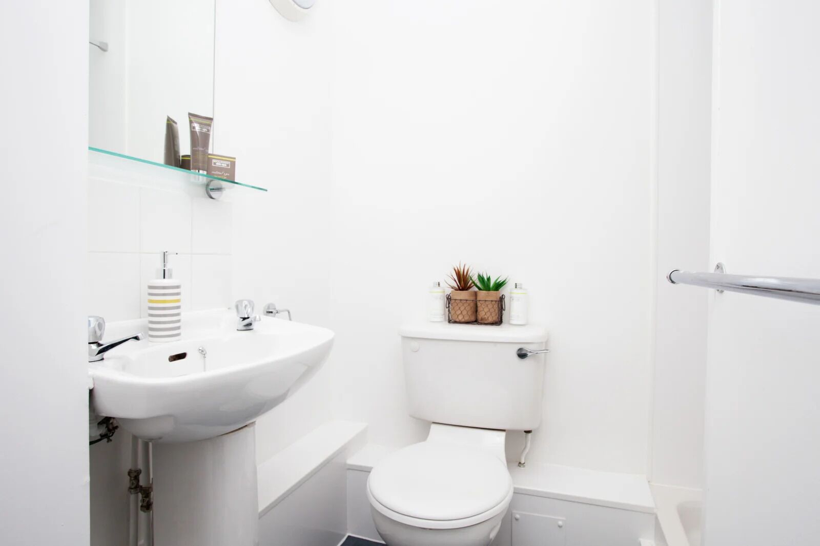 A small, white bathroom with a sink, toilet, and glass shelf holding toiletries.
