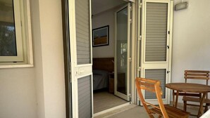 Un balcón con una mesa y sillas de madera, una puerta corredera de vidrio que da a un cuarto interior y un cuadro enmarcado en la pared.