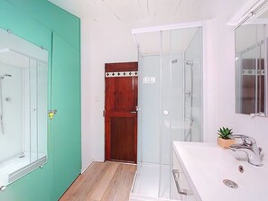 A bathroom with a glass shower enclosure, a white sink with a modern faucet, and a wooden door with a coat hook. - Le Mascoïnat by Interhome (Nice)