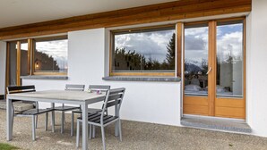 Uma área moderna de refeições ao ar livre, com mesa e cadeiras, janelas amplas com molduras de madeira e vista para árvores e montanhas.
