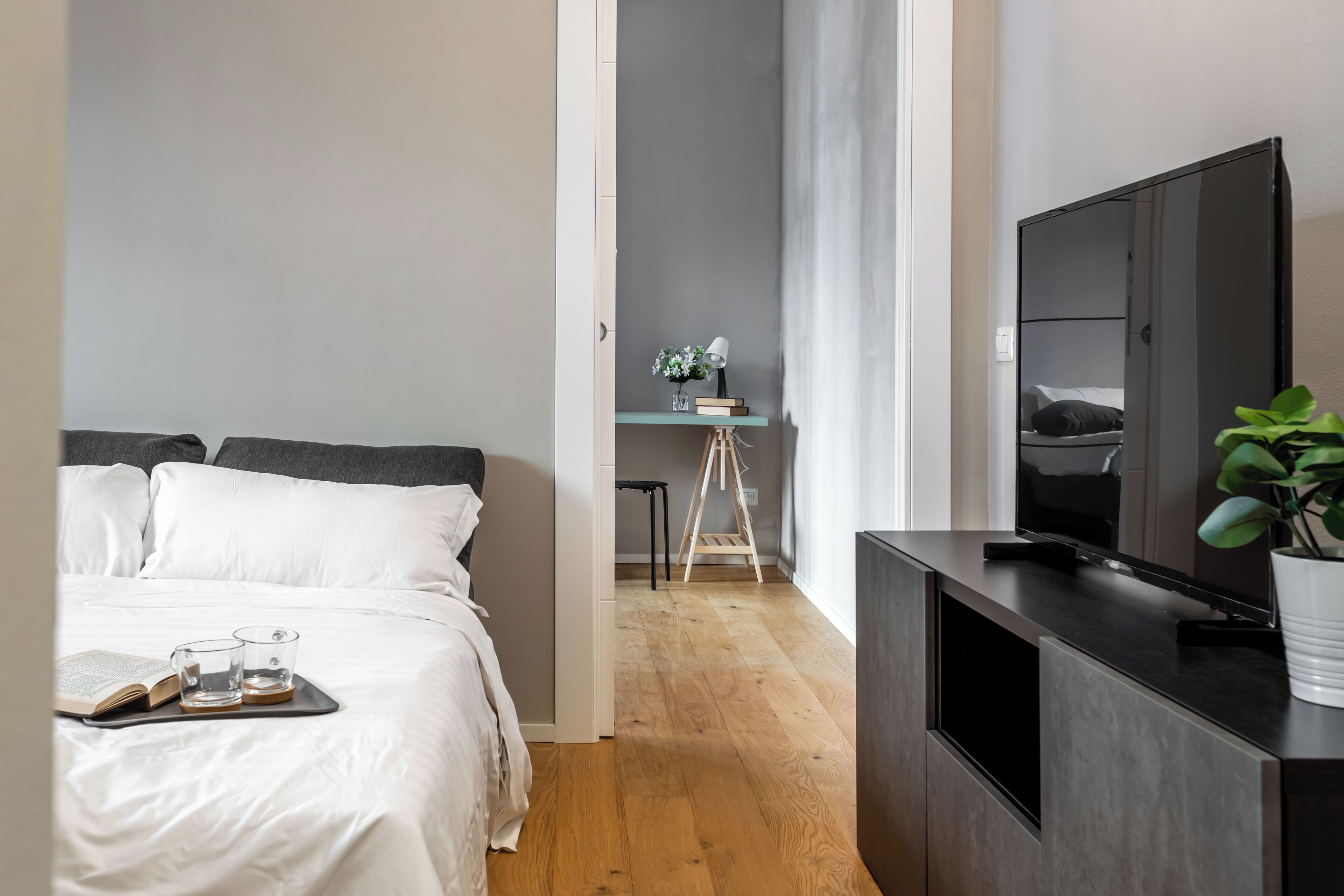 A modern hotel room with a bed, a desk with a lamp, a TV, and a plant.