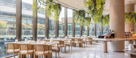 En modern restaurang med stora fönster, hängande växter och snyggt arrangerade bord och stolar.