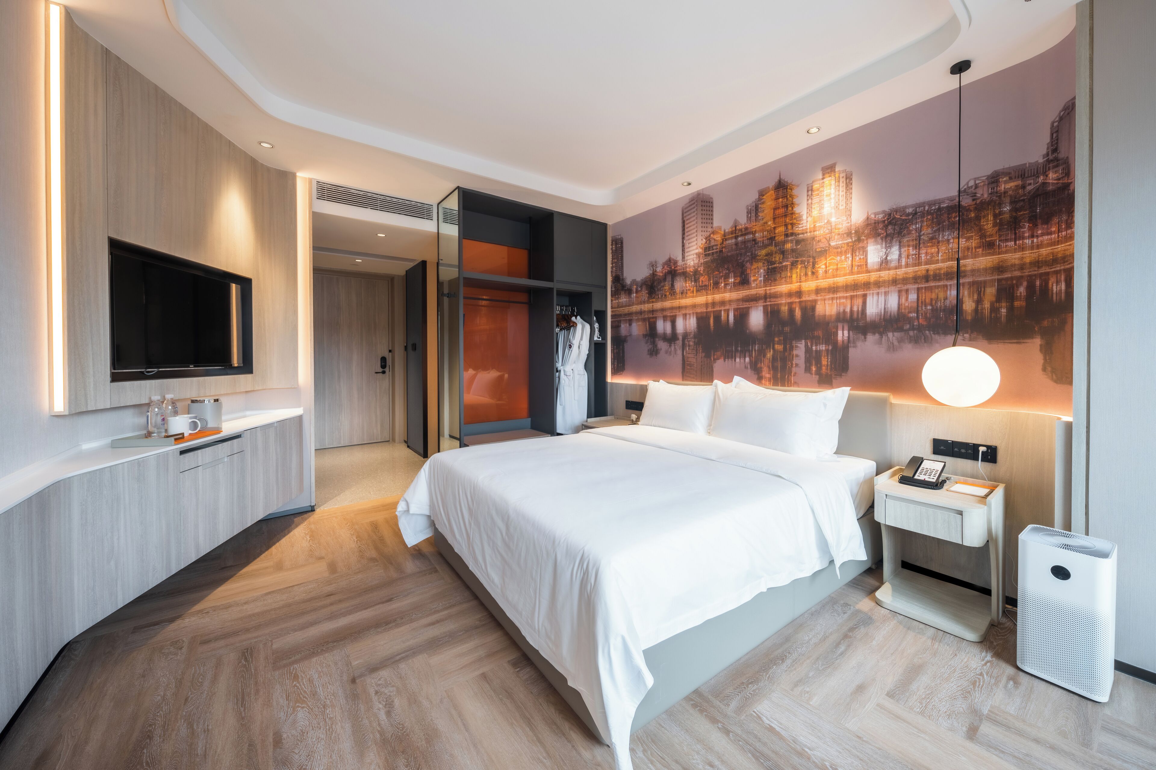 Una habitación de hotel moderna con una cama grande, un televisor de pantalla plana montado en la pared, una mesita de noche con una lámpara y un ventanal con vistas a la ciudad.