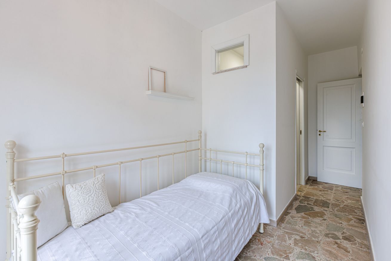 Un letto singolo con testiera e pediera bianche, un comodino piccolo e una parete spoglia con una porta e una piccola finestra.