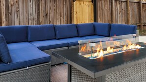 A blue sectional sofa with a glass-top fire pit in the center. - Ultimate Retreat | Game Room Fire Pit BBQ Office (San Antonio)