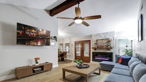 A living room with a sofa, coffee table, TV, fireplace, and wooden ceiling beams. - Ultimate Retreat | Game Room Fire Pit BBQ Office (San Antonio)