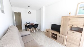A modern living room with a sofa, a TV on a wooden cabinet, and a dining area with chairs and a table. - 099 - Parque y playa 007 - comfortHOLIDAYS (Santa Pola)