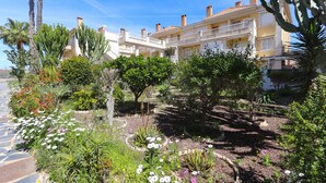 A hotel with a garden featuring various plants and flowers, a pool, and a clear blue sky. - 099 - Parque y playa 007 - comfortHOLIDAYS (Santa Pola)