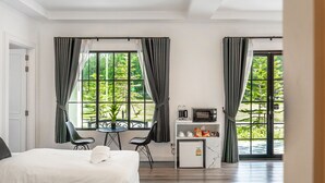 A hotel room with a bed, a desk with a chair, a microwave, a small fridge, and a view of greenery through large windows. - Tarn Villa Krabi (Krabi)