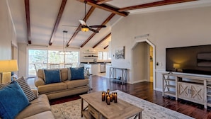 A spacious living room with a high ceiling, wooden beams, a sofa, a coffee table with candles, a TV, and a kitchen in the background. - Altitude Adjustment by Chatuge Home Concierge | Firepit, Hot Tub, Mountain & Lake Views (Hiawassee)