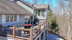 A two-story house with a deck, a covered patio, and a large window overlooking a forested area. - Altitude Adjustment by Chatuge Home Concierge | Firepit, Hot Tub, Mountain & Lake Views (Hiawassee)