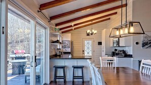 A modern kitchen with wooden beams, a large island with stools, and a dining area with a wooden table and chairs. - Altitude Adjustment by Chatuge Home Concierge | Firepit, Hot Tub, Mountain & Lake Views (Hiawassee)