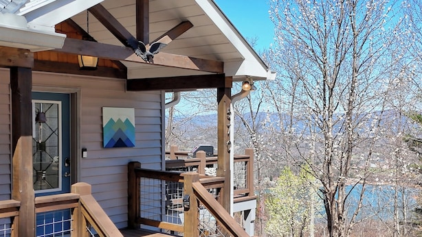 A wooden deck with a staircase leading to a covered porch area, a door with a glass panel, and a mountain view in the background. - Altitude Adjustment by Chatuge Home Concierge | Firepit, Hot Tub, Mountain & Lake Views (Hiawassee)