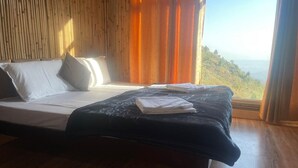 A bedroom with a bed, wooden floor, and a view of a hilly landscape through a window. - Ivory Estate Kyarkuli (Dehradun)