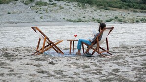 En person sidder i en strandstol med en bærbar computer, et lille bord og en drik, mens et bjergrigt landskab ses i baggrunden.