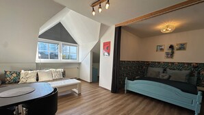 A cozy room with a loft bed, a dining area with a table and chairs, and a window with a view of buildings. - Pet-friendly one-room vacation apartment at the spa gardens (Bad Rodach)