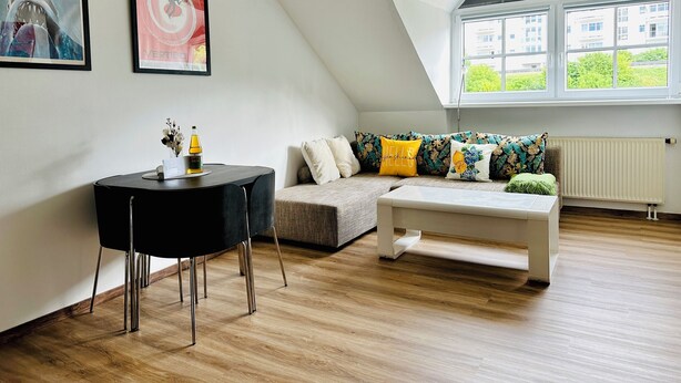A modern living room with a high ceiling, a black dining table, a sofa with colorful cushions, and a large window with a view of buildings and greenery. - Pet-friendly one-room vacation apartment at the spa gardens (Bad Rodach)