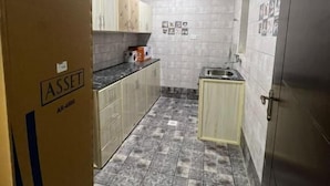 A compact kitchen with grey tiled walls, a tiled floor, and basic cabinetry. - RED3 NEW SALALAH (Salalah)